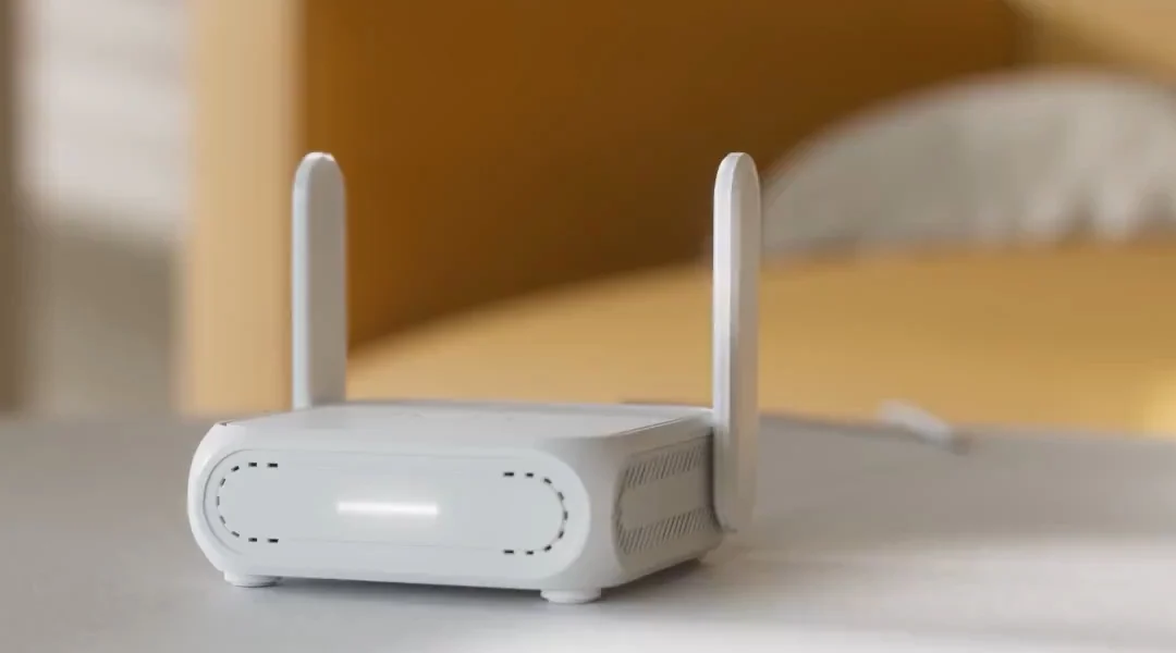 White ASUS RT-BE58 Go travel router with two upright antennas and a light bar on the front, on a bedside table